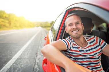 Yüzünde gülümseme olan bir adam kırmızı bir araba sürer..