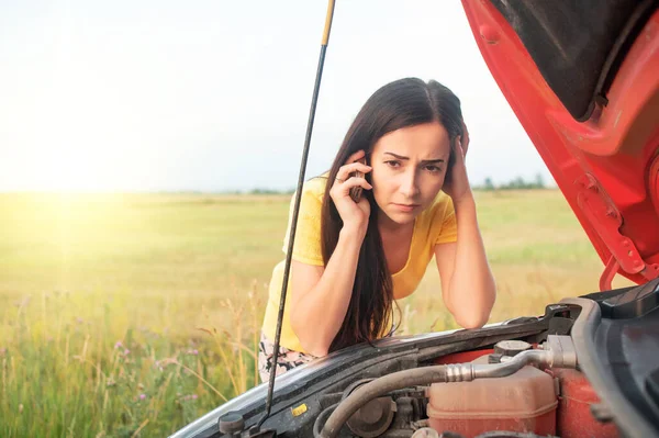 Car breakdown. Young woman calls on the phone while standing by a broken car with an open hood.