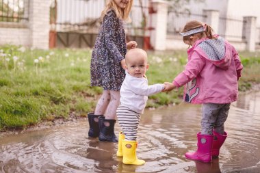 Sarı çizmeli küçük çocuk yürür. Kızlarla el ele tutuşur. Lastik botlarla su birikintisinde koşar.