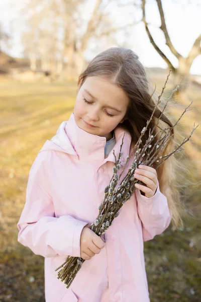 ピンクのレインコートを着た可愛い女の子春のイースターヤナギの枝を手に太陽の光を屋外で — ストック写真