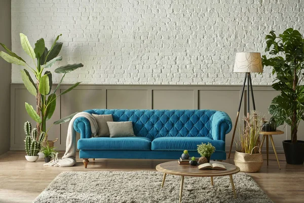 Blue quilted sofa in front of the brown classic grid and white brick wall, blanket vase of green plant lamp design, clock design.