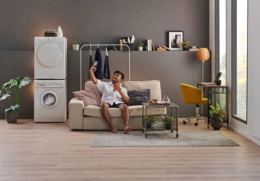 Genç adam minimalist ev tarzında yaşıyor, laptopta çalışıyor, çamaşır makinesi, çamaşır makinesi, odada telefona bakıyor, gri duvar arka planı ve dekoratif iç mekan.