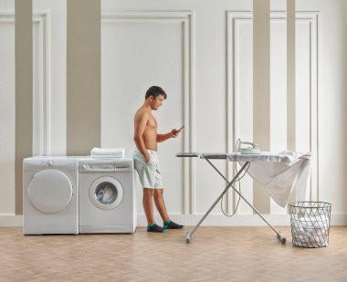 Adam banyoda ütü yapıyor, çamaşır ve kurutma makinesi beyaz ve kahverengi duvarların önünde..