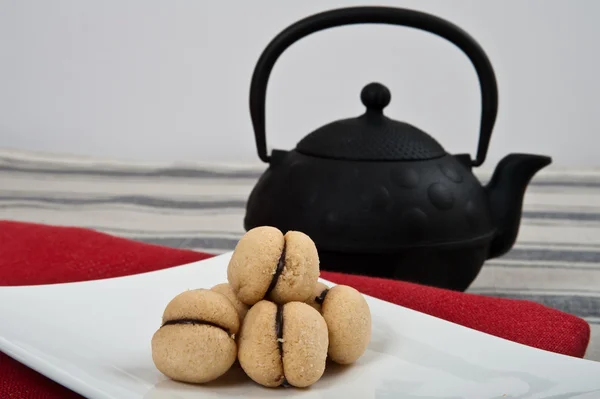 Biscoitos italianos chamados "baci di dama" em uma placa branca com um pano vermelho e um grande bule preto . — Fotografia de Stock