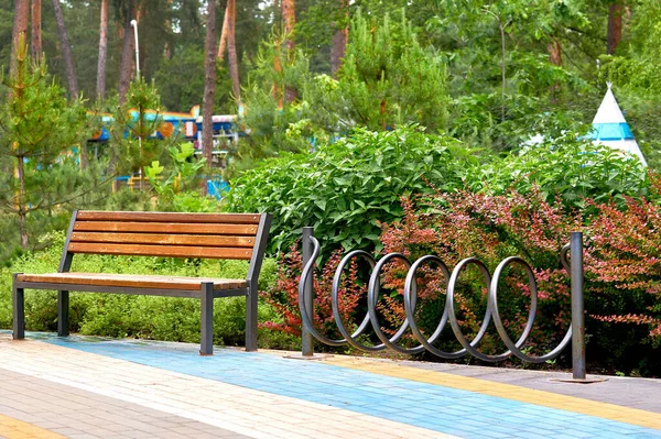 a large public green area in a town, used for recreation. Wooden bench and bike rack in a green city park with rides and trees