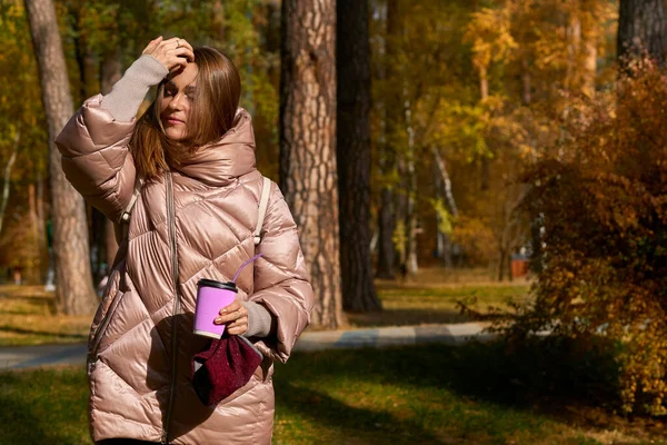 an adult female human being. Pretty young woman in a pink jacket straightens her hair with a glass of coffee tea in the autumn park