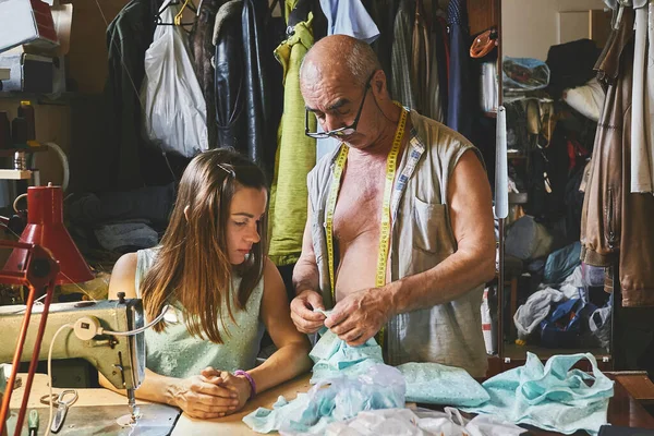 a person whose occupation is making fitted clothes such as suits, pants, and jackets to fit individual customers. Senior experienced master tailor teaches a young girl seamstress how to sew