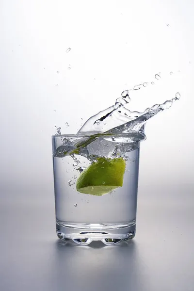 Pedaço Limão Salpicando Copo Líquido Claro Água Uma Superfície Cinza — Fotografia de Stock