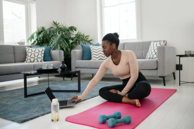 Siyah Afrikalı kadın dizüstü bilgisayarla spor yapıyor. Yoga minderinde oturmak, evdeki modern oturma odasının konforunda dambılları tutmak.