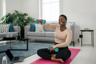 Bilgisayarda spor yaptıktan sonra gülümseyen siyahi Afrikalı kadın. Yoga minderinde oturmak, evdeki modern oturma odasının konforunda dambılları tutmak.