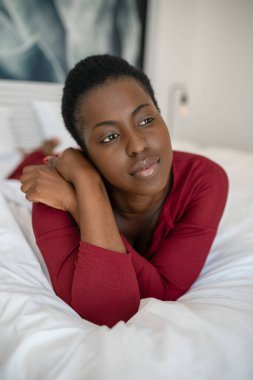Güzel siyah Afrikalı kadın portresini yakından çek. Yatakta uzanmış, kollarını kavuşturmuş, başka tarafa bakıyor.