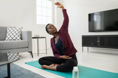 Siyahi Afrikalı bir kadının yoga pozisyonunda oturma odasında mindere oturması, esnemesi ve evde meditasyon yapması.