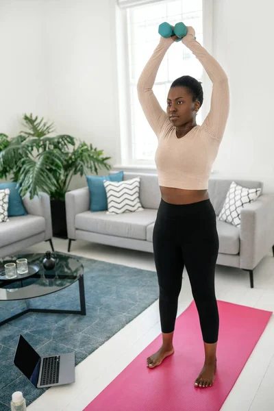 Black African woman exercising at home. Doing weighted dumbbell workout, arms raised in modern living room from home