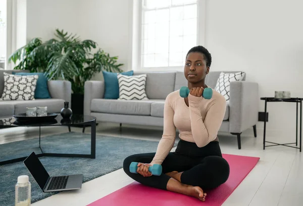 Black African woman following online fitness workout on laptop. Sitting on yoga mat, holding dumbbells in the comfort of modern living room at home.