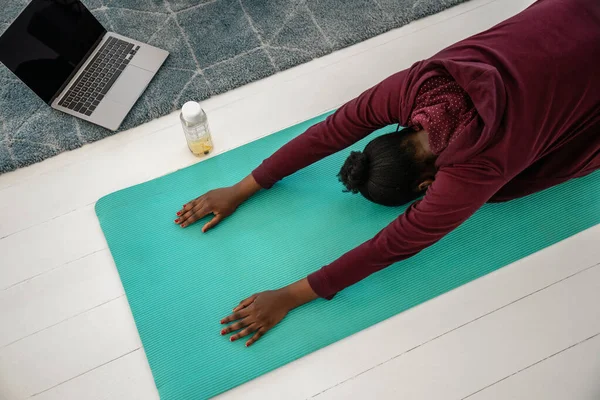 Young Black African woman doing yoga exercises online. Stretching at home on mat in front of laptop computer, shot directly from above.