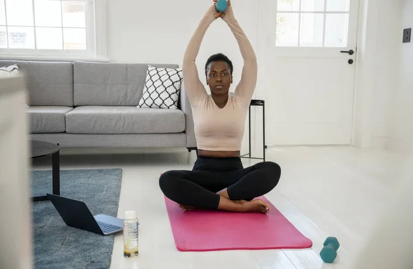 Black African woman following online fitness workout on laptop. Sitting on yoga mat, holding dumbbells in the comfort of modern living room at home