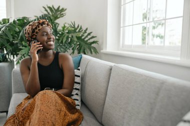 Kanepede oturan, elinde akıllı bir telefonla gülümseyen, gülen ve fotokopi çeken güzel bir Afrikalı kadın.