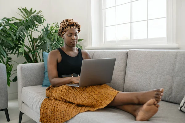 Beautiful traditional young Black gen z African woman sitting on sofa at home, typing on laptop, smiling, shot with copy space