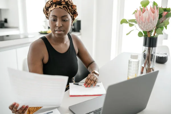 Black African woman working from home, going through paperwork and writing down notes. Laptop and smartphone on home office table