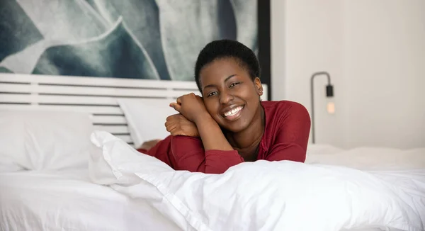 Panoramic portrait of beautiful African woman laying on bed with legs and arms crossed, smiling and looking into the camera with copy space