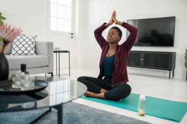 Siyahi Afrikalı kadın yoga pozisyonunda oturma odasında minder üzerinde oturuyor, evde egzersiz yapıyor ve meditasyon yapıyor.