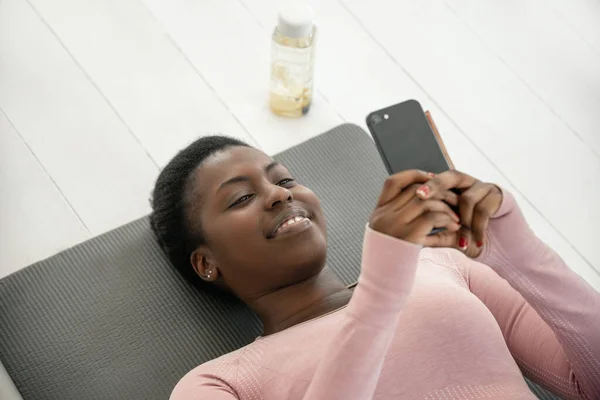 Black African woman laying on yoga mat smiling and looking at phone, resting and taking a break from exercising. shot from above with copy space