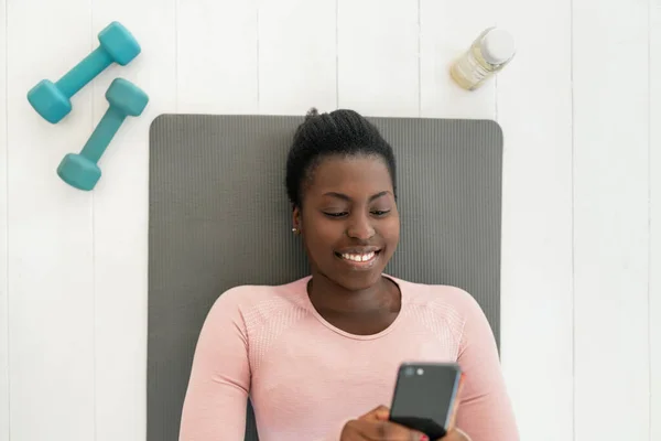 Black African woman laying on yoga mat smiling and looking at phone, resting and taking a break from exercising. Shot from above with copy space
