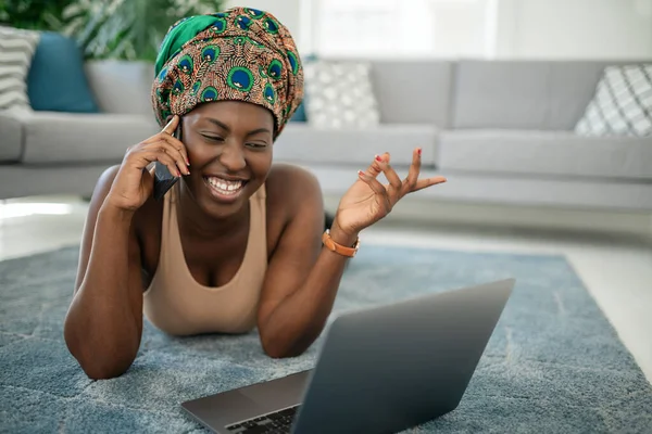 Beautiful African woman at home, smiling and laughing, using mobile smart phone, wearing traditional headscarf, surprised and happy