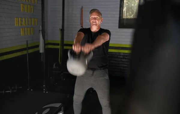 Mature man exercising in home garage gym. Senior man doing kettlebell swing exercise, showing motion.
