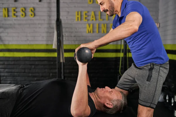 Side view of personal trainer assisting mature man in doing weighted seated arm curls at indoor garage home gym, strength training for elderly man