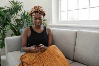 Beautiful traditional young Black gen z African woman sitting on sofa at home, typing on smart phone, smiling, shot with copy space
