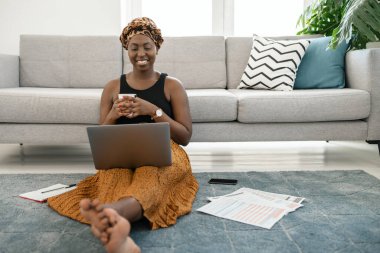 Beautiful black African woman sitting on floor in lounge, work meeting on laptop at home with coffee in hand wearing traditional head scarf