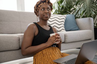 Beautiful young gen z Black African woman, wearing traditional head scarf, sitting on lounge floor holding coffee cup and laptop on lap