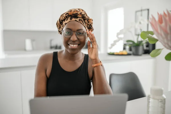 Black African woman smiling, holding eye glasses. Looking at laptop while sitting and working from home. Wearing traditional head scarf