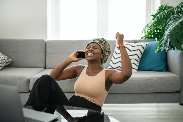 Beautiful Black African woman at home, smiling and laughing, using mobile smart phone, wearing traditional headscarf, surprised and happy, fist in air