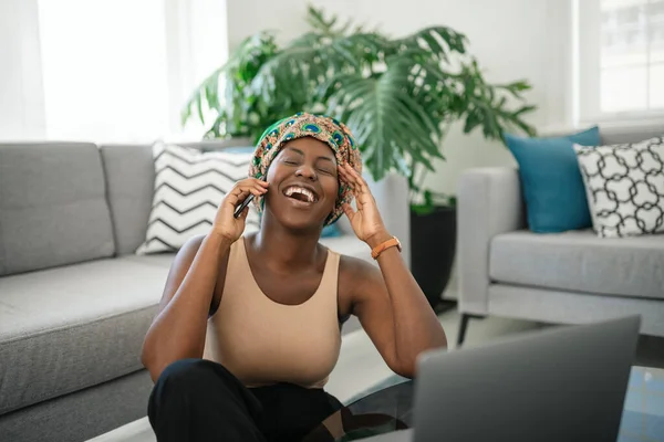 Beautiful Black African woman at home, smiling and laughing, using mobile smart phone, wearing traditional headscarf, surprised and happy