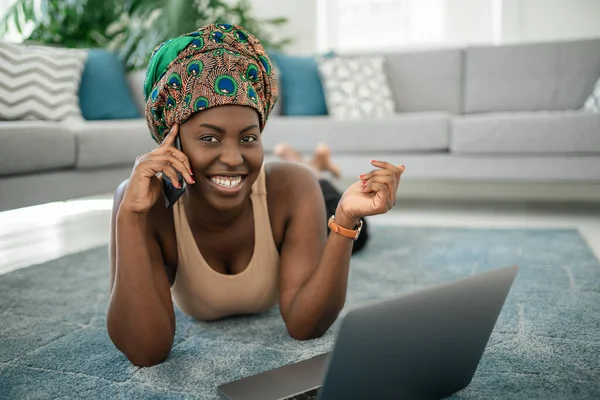 Beautiful African woman at home, smiling and laughing looking into camera, using mobile smart phone, wearing traditional headscarf, surprised and happy