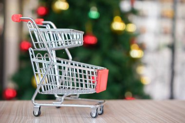 Yeşil Noel ağacında boş bir sepetin içinde kırmızı alışveriş arabası ve mevsim kutlaması. İnternetten tatil alışverişi. Ahşap masada küçük bir tramvay.