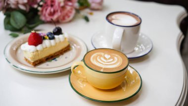 Kahve dükkanındaki beyaz seramik bardağın üstünde lale sütlü sütlü latte ile Latte sanat kahvesi. Tatlı menüsü olarak New York pastası ve kısa bardakta beyaz köpüklü sıcak çikolata içeceği..