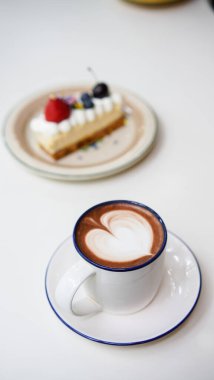 Kahve dükkanındaki beyaz seramik bardağın üstünde lale sütlü sütlü latte ile Latte sanat kahvesi. Tatlı menüsü olarak New York pastası ve kısa bardakta beyaz köpüklü sıcak çikolata içeceği..