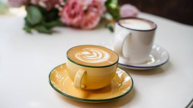 Kahve dükkanındaki beyaz seramik bardağın üstünde lale sütlü sütlü latte ile Latte sanat kahvesi. Tatlı menüsü olarak New York pastası ve kısa bardakta beyaz köpüklü sıcak çikolata içeceği..
