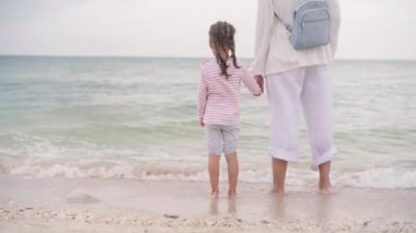 Anne, küçük kızıyla birlikte sahilde mutlu bir ailede yürüyor. Anne, deniz kıyısında küçük bir çocuğun elini tutuyor. Seyahat Yaz Tatili 