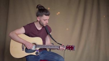 Akustik gitar çalan yakışıklı genç adam. Oturma odası kumaş arka planda.