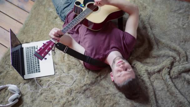 Homme avec guitare acoustique chantant et jouant de la guitare couché par terre près d'un ordinateur portable et d'écouteurs. Vue de dessus. Évasion de l'ennui. — Video