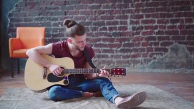 Akustik gitar çalan yakışıklı genç adam oturma odasında oturuyor.