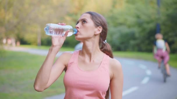 Vrouwelijke loper drinken water transparant plastic fles na ochtend joggen Kaukasische atletische vrouwelijke rusten na training zomerdag in het park — Stockvideo