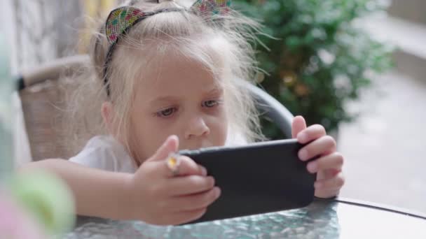 Девочка смотрит видео на экране смартфона сидя под открытым небом кафе — стоковое видео