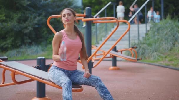 辛勤训练后，瘦弱的女人坐在运动场上休息，喝着水，喝着塑料瓶，打了一针 — 图库视频影像