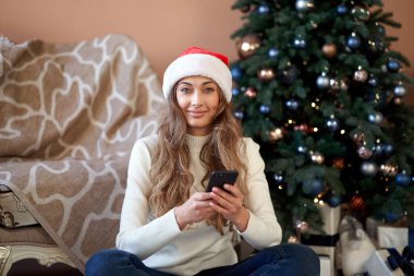 Noel kadını beyaz kazak giymiş Noel Baba şapkası takıyor. Beyaz Noel ağacının yanında oturuyor. Kafkasyalı kadın kış tatilini rahatlatmak için akıllı telefon internet alışveriş sezonu kullanıyor. 