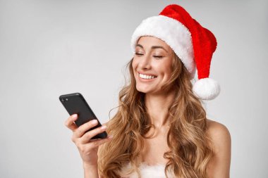 Noel Baba Beyaz Şapkalı Kadın stüdyosunun arka planında elinde akıllı telefon güzel beyaz kadın kıvırcık saçlı portresi var. Mutlu insan pozitif duygu. Fotokopi alanı ile tatil kavramı. Gülümseyen diş. 